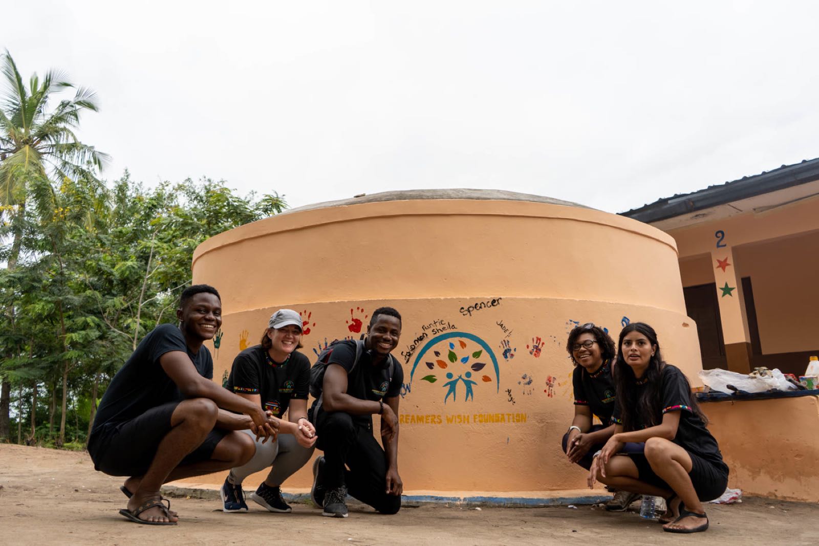 Founding volunteers squatting in front of the Dreamers Wish water tank in Dadome, covered in children's handprints and the names Sheila, Jade and Angie